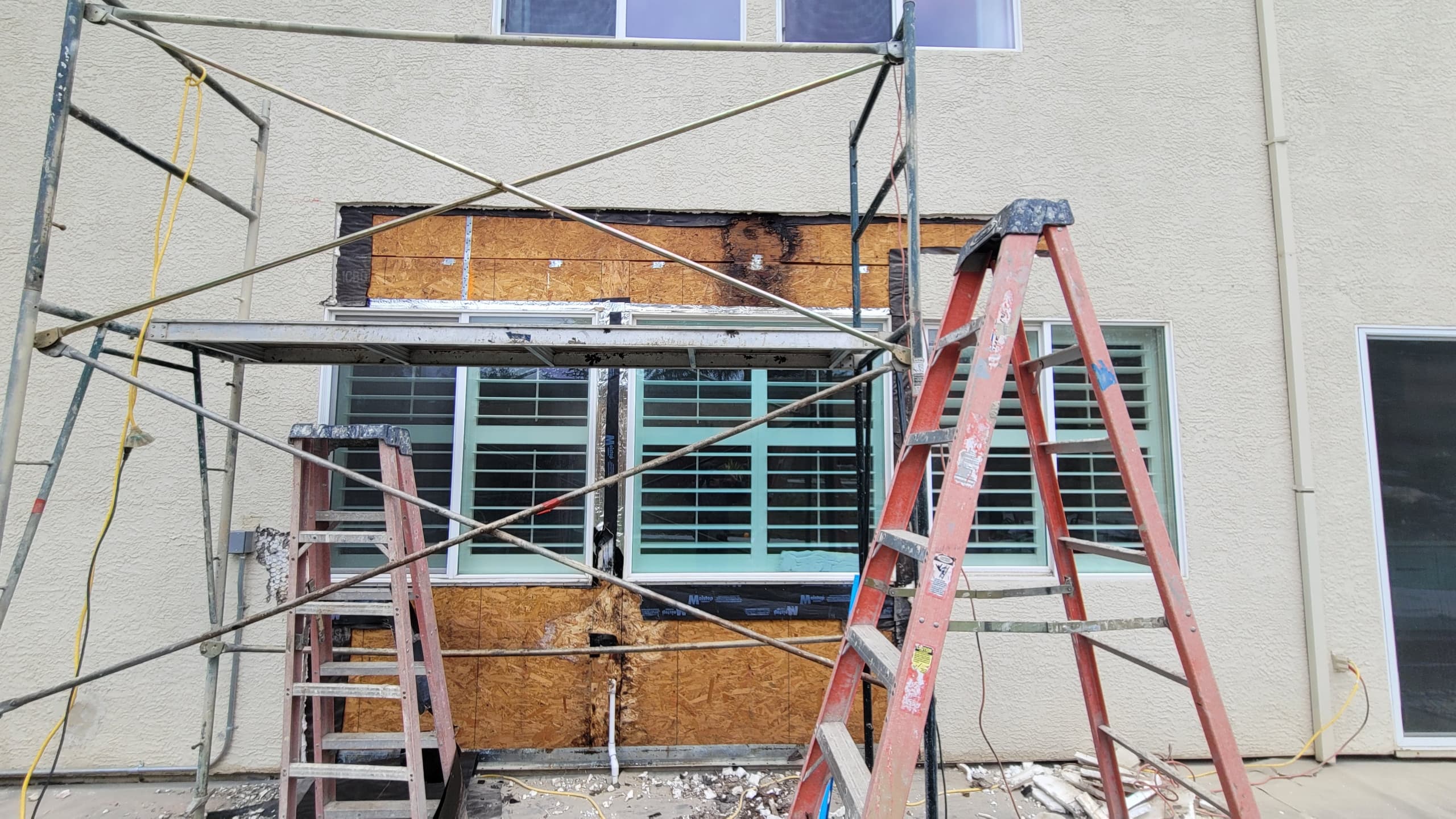 Scaffolding and orange ladders are positioned against a stucco house where a rectangular section of stucco has been removed below an upper window, revealing dark-stained plywood sh